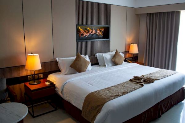 The image shows a modern hotel room with a large bed, decorative cushions, bedside lamps, and a wall art above the bed.