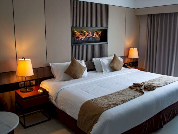 The image shows a modern hotel room with a large bed, decorative cushions, bedside lamps, and a wall art above the bed.