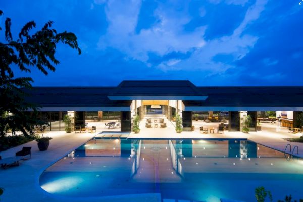The image shows a luxurious building with a large swimming pool, surrounded by a well-lit area under a twilight sky.