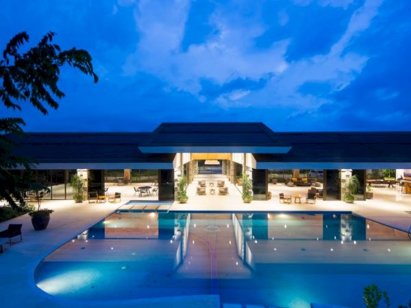 The image shows a luxurious building with a large swimming pool, surrounded by a well-lit area under a twilight sky.