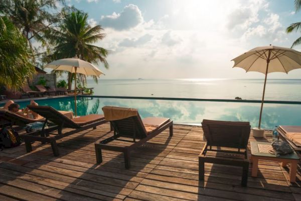 A serene poolside scene with wooden deck chairs, umbrellas, and a view of palm trees and the ocean under a partly cloudy sky.
