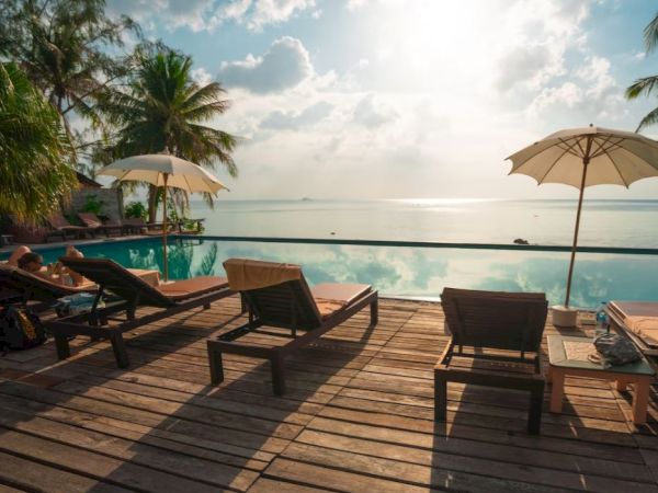 A serene poolside scene with wooden deck chairs, umbrellas, and a view of palm trees and the ocean under a partly cloudy sky.