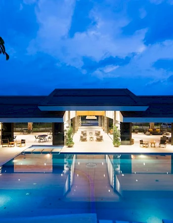 The image shows a luxurious modern building with large glass windows, surrounded by a well-lit swimming pool under a cloudy evening sky.