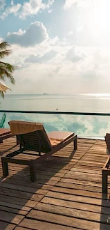 A serene poolside scene with lounge chairs, umbrellas, palm trees, and a calm ocean under a cloudy sky.