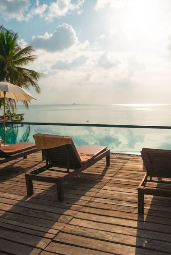 A serene beachfront with lounge chairs, umbrellas, and palm trees by the pool overlooking the ocean under a partly cloudy sky.