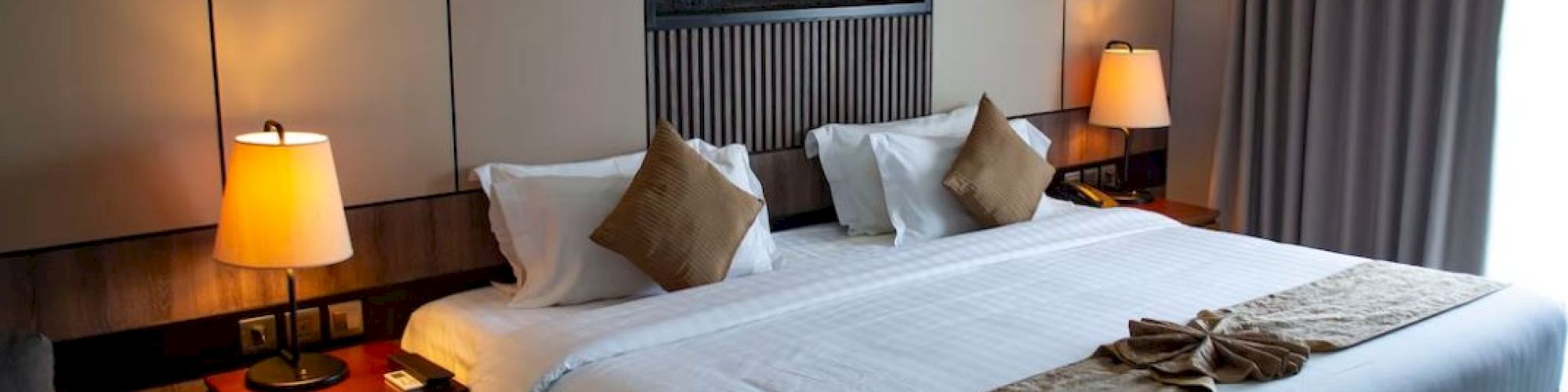 A cozy hotel room with a large bed, bedside tables, lamps, and a window with curtains. There’s a decorative piece above the bed.
