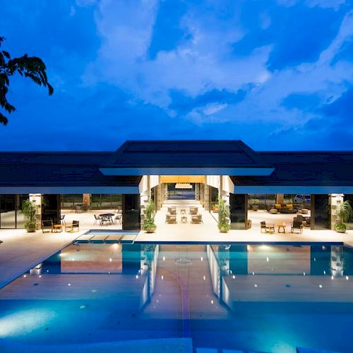 Luxurious outdoor pool area at dusk, featuring modern architecture and ambient lighting, surrounded by greenery and open seating.