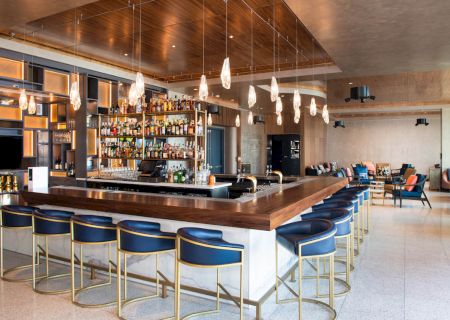 The image shows a modern bar with a wooden counter, blue chairs, hanging lights, and a well-stocked shelf of bottles, within a stylish space.