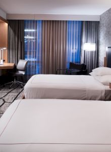 The image shows a modern hotel room with two beds, a large TV, a desk with a chair, stylish lighting, patterned carpet, and wall decor.