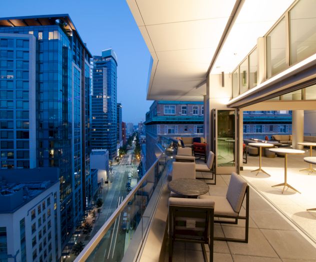 A modern cityscape at dusk features a balcony with tables and chairs, overlooking tall buildings and illuminated streets below.