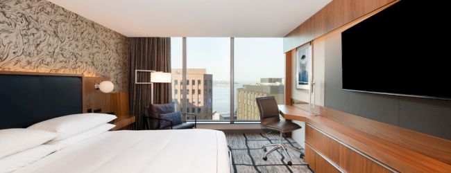 A modern hotel room features a large bed, a wall-mounted TV, a desk, and a window with city views.