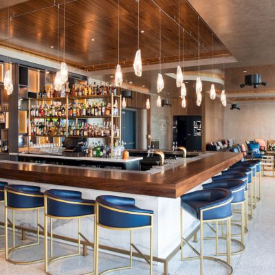 This image shows a modern bar with hanging lights, a stocked liquor display, blue bar stools, and a seating area in the background.
