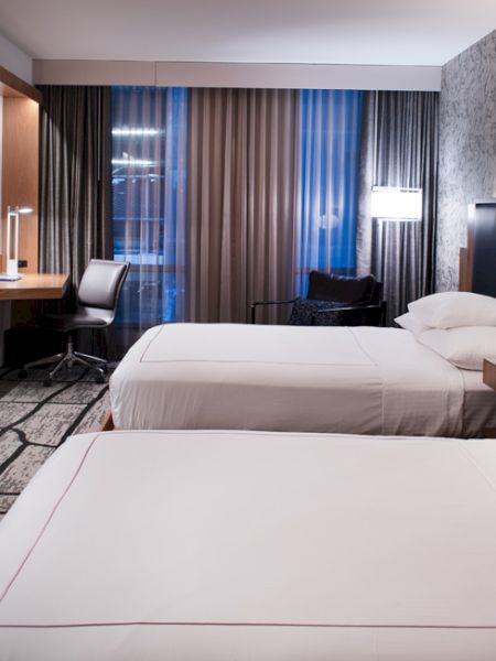 A modern hotel room with two beds, a desk, and a chair. The decor is sleek with a neutral color palette.