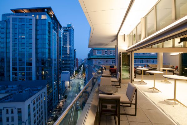 A modern balcony overlooking a city street with tall buildings, featuring outdoor seating and tables, illuminated in the evening.
