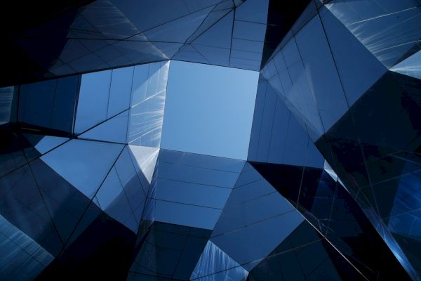 Abstract view of a modern building's reflective glass facade, looking upwards towards a clear blue sky through angular structures.