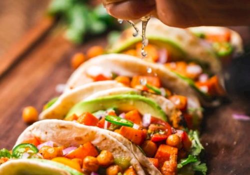 The image shows tacos filled with chickpeas, vegetables, and avocado, with lime being squeezed over them on a wooden board.