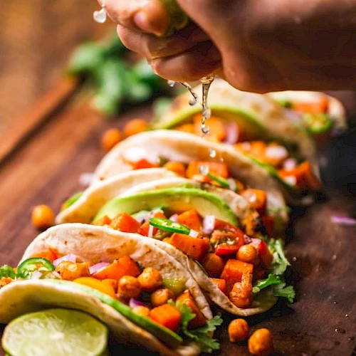 The image shows tacos filled with chickpeas, vegetables, and avocado, with lime being squeezed over them on a wooden board.