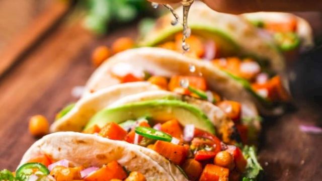 The image shows tacos filled with chickpeas, vegetables, and avocado, with lime being squeezed over them on a wooden board.