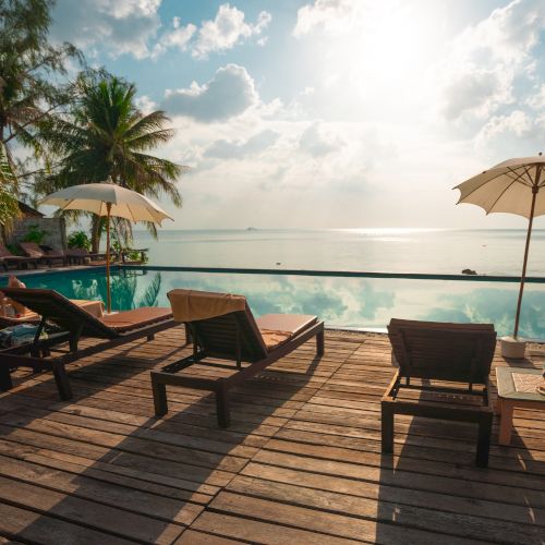 A tropical setting with sun loungers and umbrellas by a pool, overlooking the ocean under a partly cloudy sky.