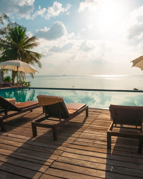 A tropical setting with sun loungers and umbrellas by a pool, overlooking the ocean under a partly cloudy sky.