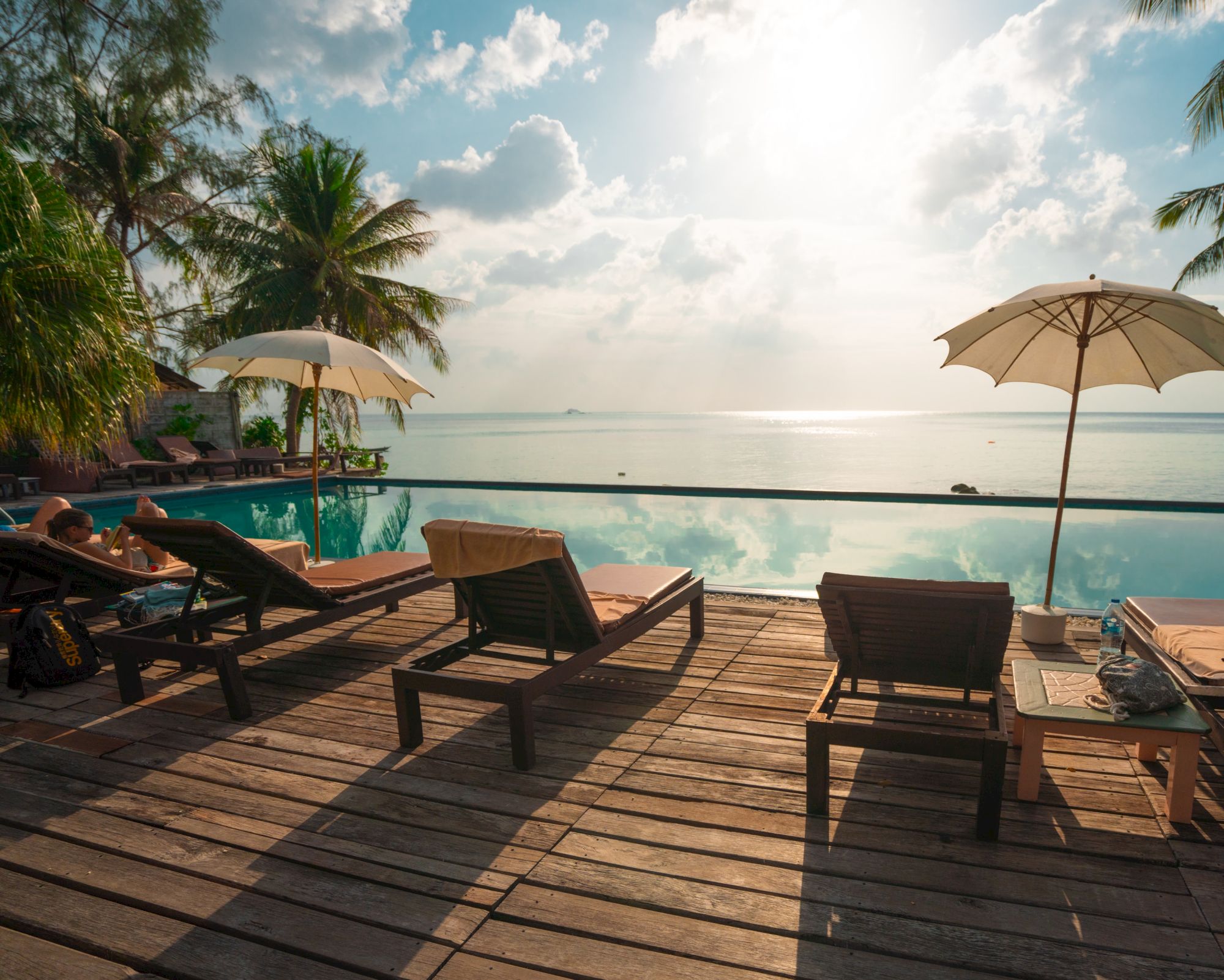 A serene beach scene with sun loungers, umbrellas, and palm trees overlooking a calm ocean under a partly cloudy sky.