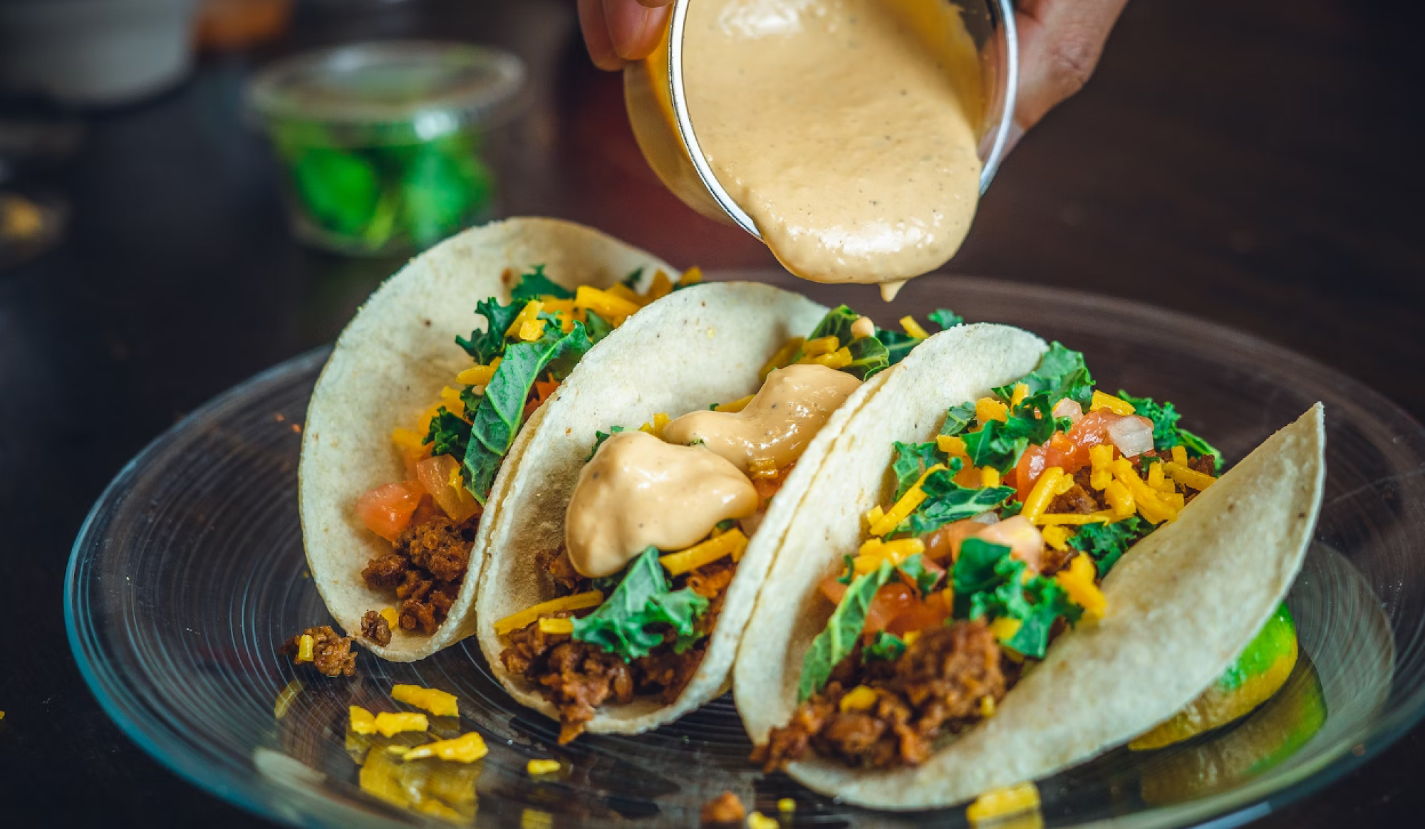 Three tacos with meat, cheese, kale, and tomatoes on a plate; sauce is being poured on top of them.