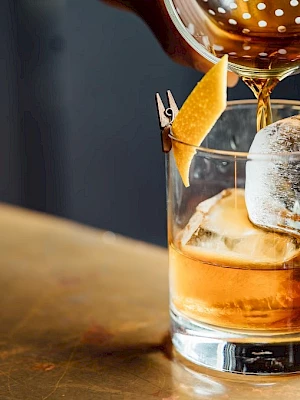 A hand pours a cocktail into a glass with ice and a citrus peel garnish on a wooden surface.