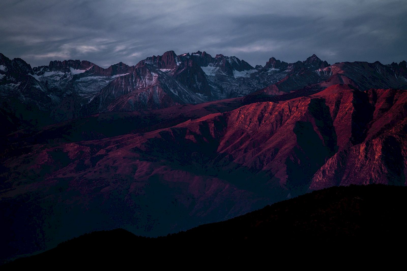 A mountain range with a dramatic sky, illuminated by sunset hues casting vibrant red and purple tones over the rugged terrain.