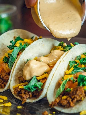 Three tacos filled with meat, cheese, tomato, and greens, being topped with creamy sauce.