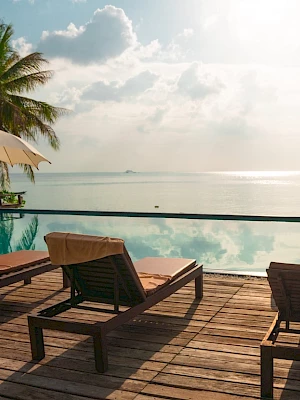A serene beach setting with lounge chairs, umbrellas, a pool, and palm trees overlooking a calm sea under a cloudy sky.