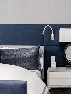 A stylish bedroom with a dark blue headboard, pillows, and a side table featuring a decorative lamp and a wall-mounted reading light.