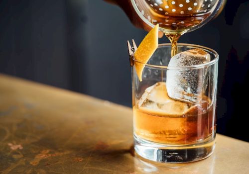 A cocktail is being poured into a glass with a large ice cube and an orange peel garnish clipped to the rim, on a bar counter.
