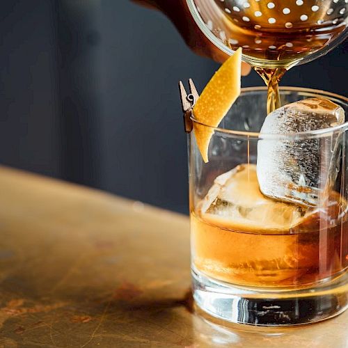 A cocktail is being poured into a glass with a large ice cube and an orange peel garnish clipped to the rim, on a bar counter.