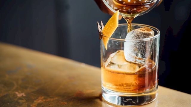 A cocktail is being poured into a glass with a large ice cube and an orange peel garnish clipped to the rim, on a bar counter.