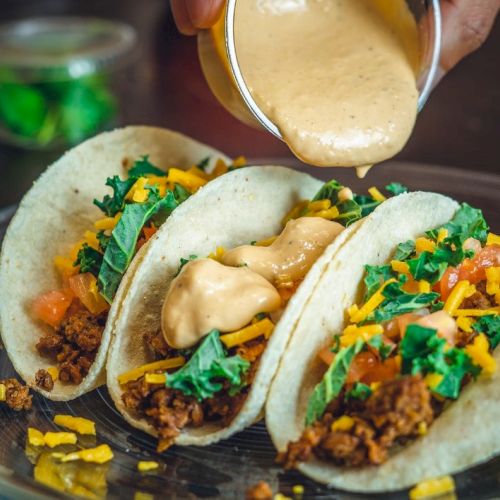 Three tacos filled with ground meat, shredded cheese, lettuce, and tomato are being topped with a creamy sauce on a plate.