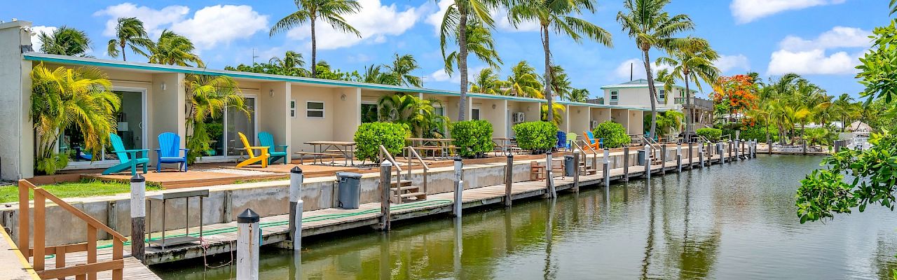 Waterfront scene with small houses, colorful chairs, palm trees, and a wooden dock beside calm water under a blue sky with clouds.