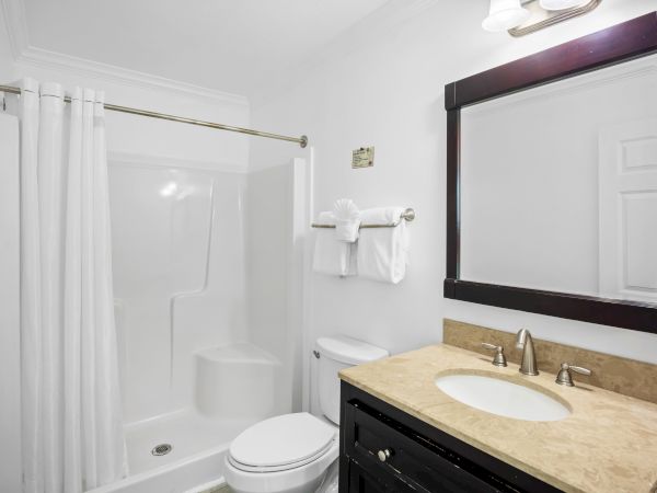 The image shows a clean bathroom with a shower, toilet, and sink. A mirror and towels are also present.