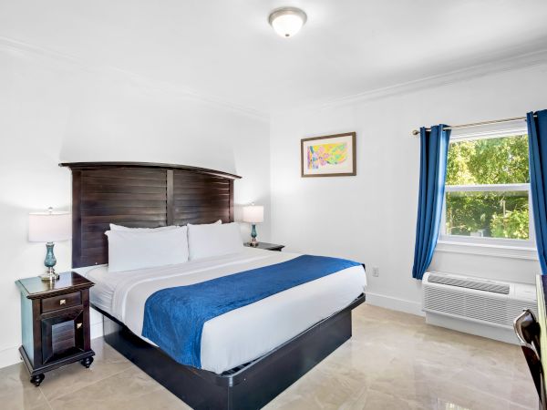 A tidy bedroom with a double bed, blue accents, two side tables with lamps, a wall painting, and a window with blue curtains.