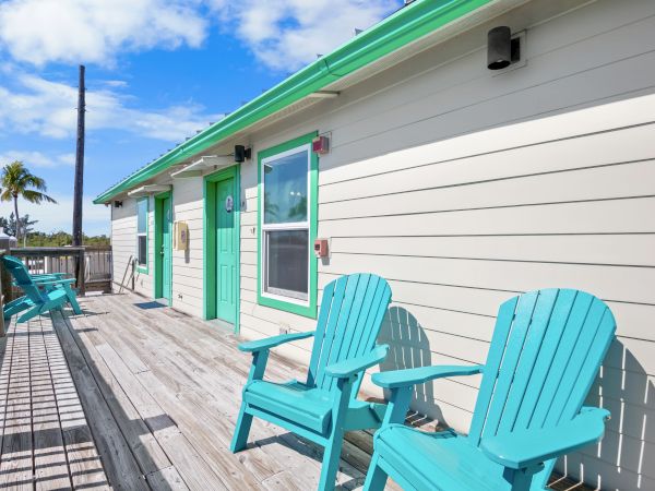 The image shows a beach house with turquoise chairs on a wooden deck, set against a sunny sky with clouds and a palm tree.