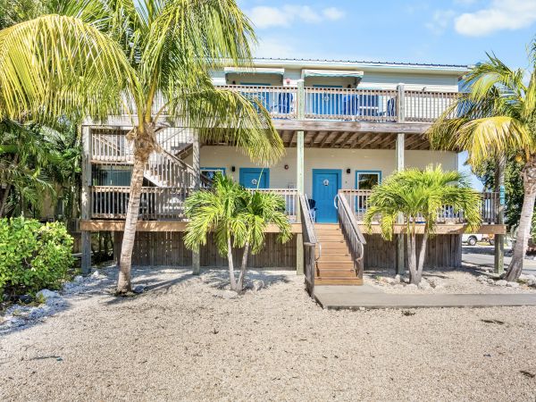 A two-story beach house with palm trees, wooden railings, and sandy ground in the front.
