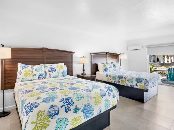 The image shows a bright, modern hotel room with two beds featuring colorful floral bedding and bedside lamps, with a view outside.