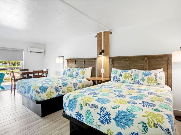 The image shows a modern hotel room with two beds, vibrant bedding, a small dining table, and a view of outdoor seating through a window.