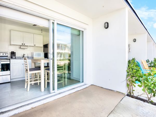 A patio with sliding doors opens to a kitchen with white cabinets, a table, and a stove; two colored chairs are outside.