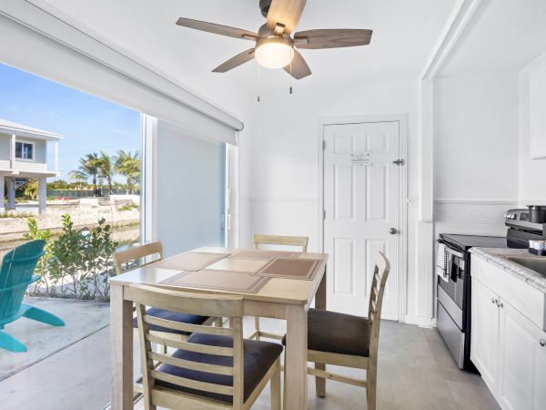 A bright kitchen with a table, chairs, ceiling fan, appliances, and a view of a patio with chairs, greenery, and a house outside.