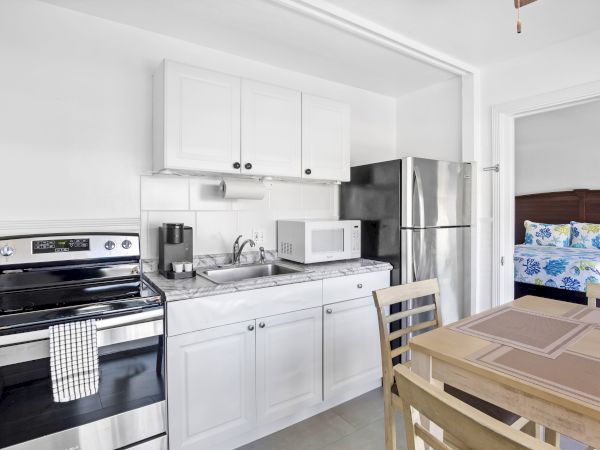 A small kitchen with a stove, fridge, microwave, coffee maker, and dining table, next to a bedroom with floral bed sheets.