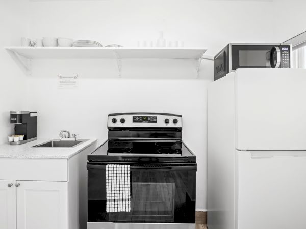 A kitchen with a stove, refrigerator, microwave, sink, coffee maker, and open shelf with dishes and cups.