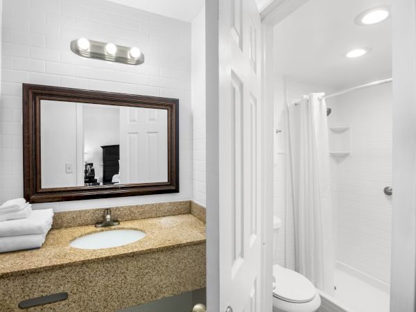 A bathroom with a granite countertop, mirror, stacked towels, sink, and adjoining shower area with a curtain and toilet.