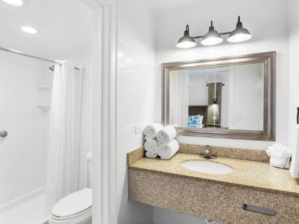 A bathroom with a granite sink, mirror, rolled towels, and overhead lights; shower and toilet are visible in the background.