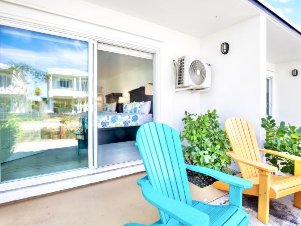A patio with colorful chairs overlooks a sliding glass door leading to a bright bedroom. Lush greenery adds a fresh outdoor touch.