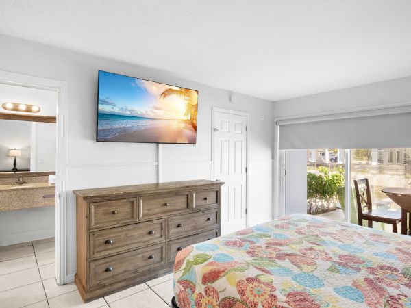 A bedroom with a colorful bedspread, wooden dresser, wall-mounted TV, and a view of a table outside.